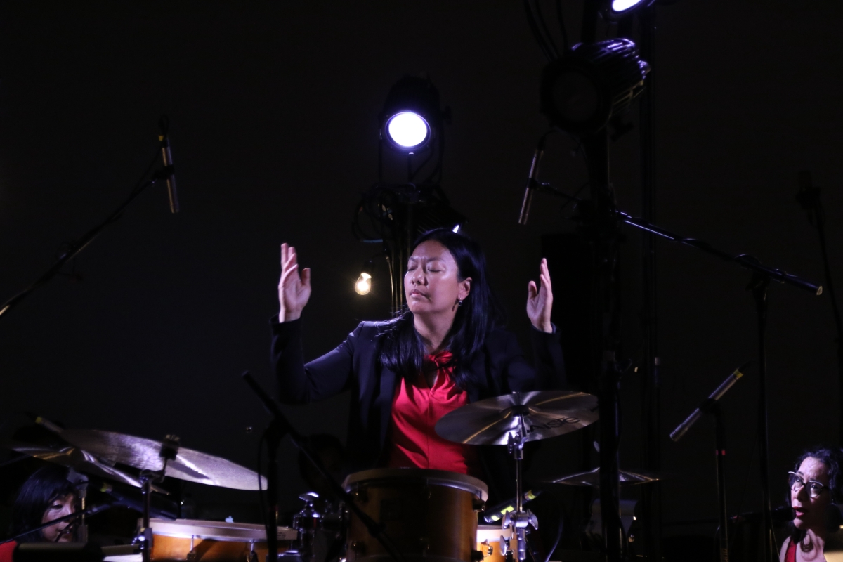 Drummer and composer Susie Ibarra conducting. She is wearing a red top and black blazer. 