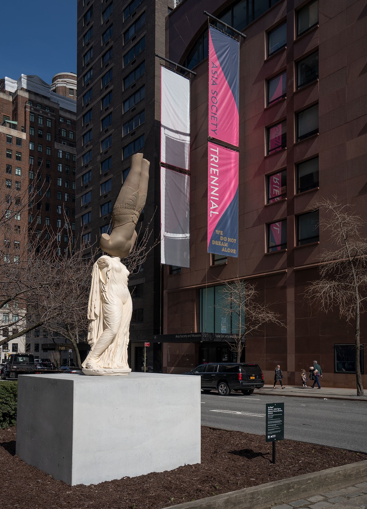 A plinth appears on a median outside of Asia Society Museum. On it is a sculpture composed of two casts: on top an inverted bronze-colored eleventh-century male figure from Cambodia, balanced atop a light beige cast of a second-century Roman figure of Venus Genetrix. Neither figure has a head.
