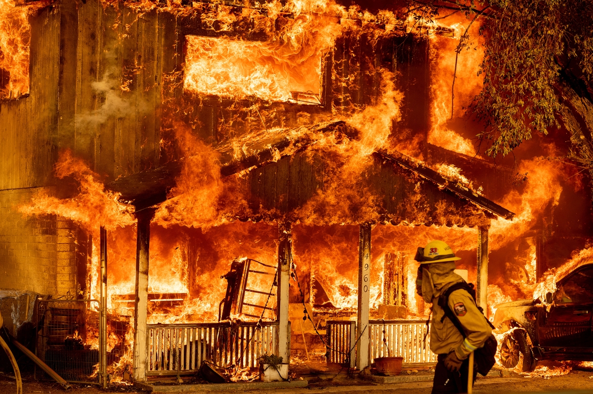 A color photograph of a house engulfed in flames. A firefighter in profile observes the inferno..