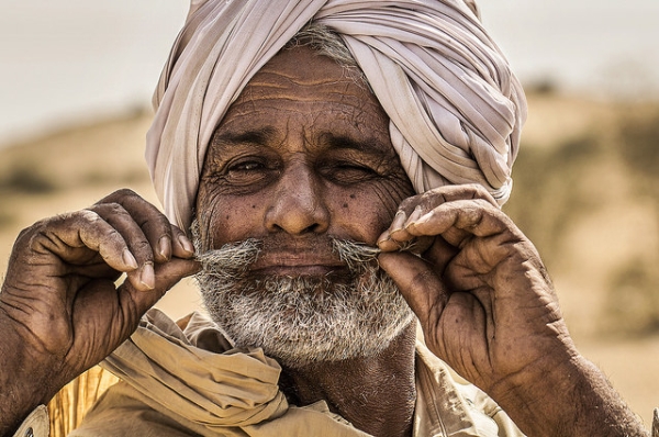 An elderly man twirls his mustache while winking at the camera in Jaisalmer, India on March 27, 2015. (Timo/Flickr)