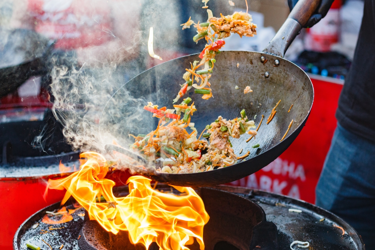 dynamic picture of a wok in motion, stir-frying colourful vegetables and meat. There's a bright flame contrasting the colour of the wok; thick smoke partially covering the wok and the dish, hinting at the rich smoked flavour the dish will have 
