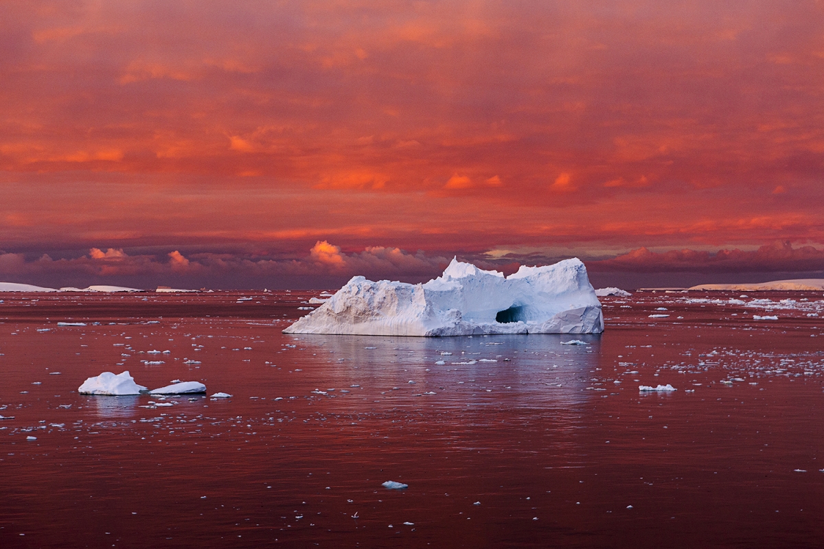 A red-orange sky is reflected in the sea. A lone large ice floe/ice berg is in the center of the image.