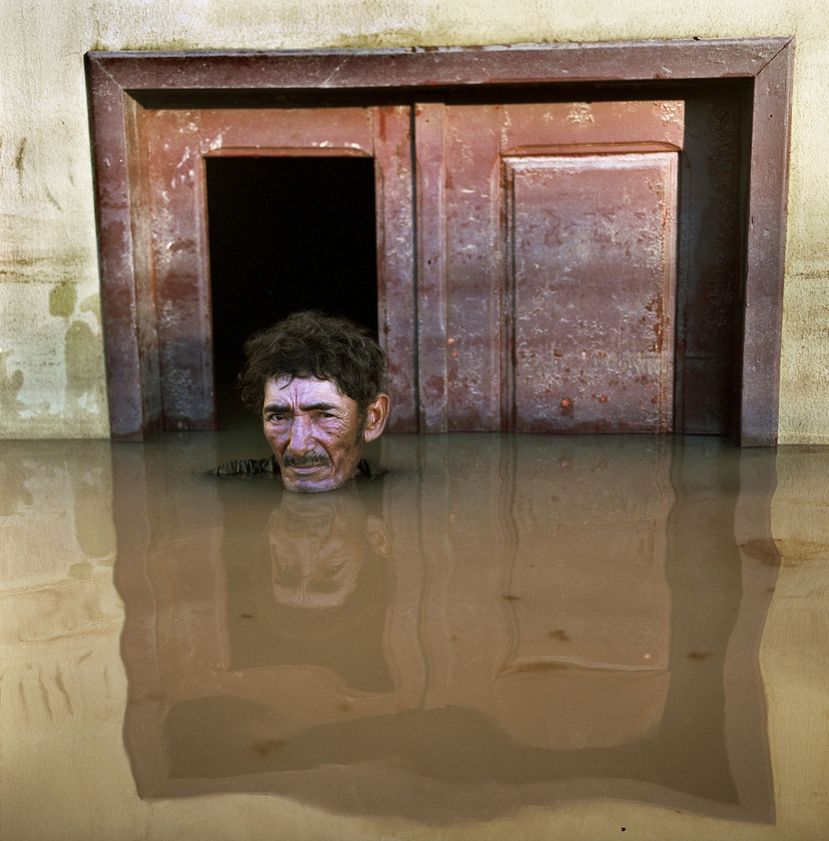 A color photograph of a man submerged up to his chin in brackish water in front of a wooden doorway. 