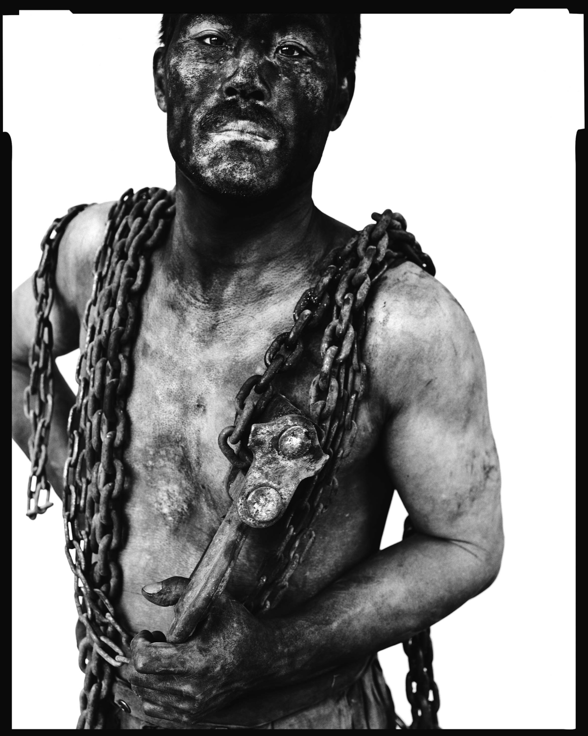 Black-and-white photograph of a three-quarter view of a bare-chested man covered in soot holding a large tool; heavy chains are draped over his shoulders.