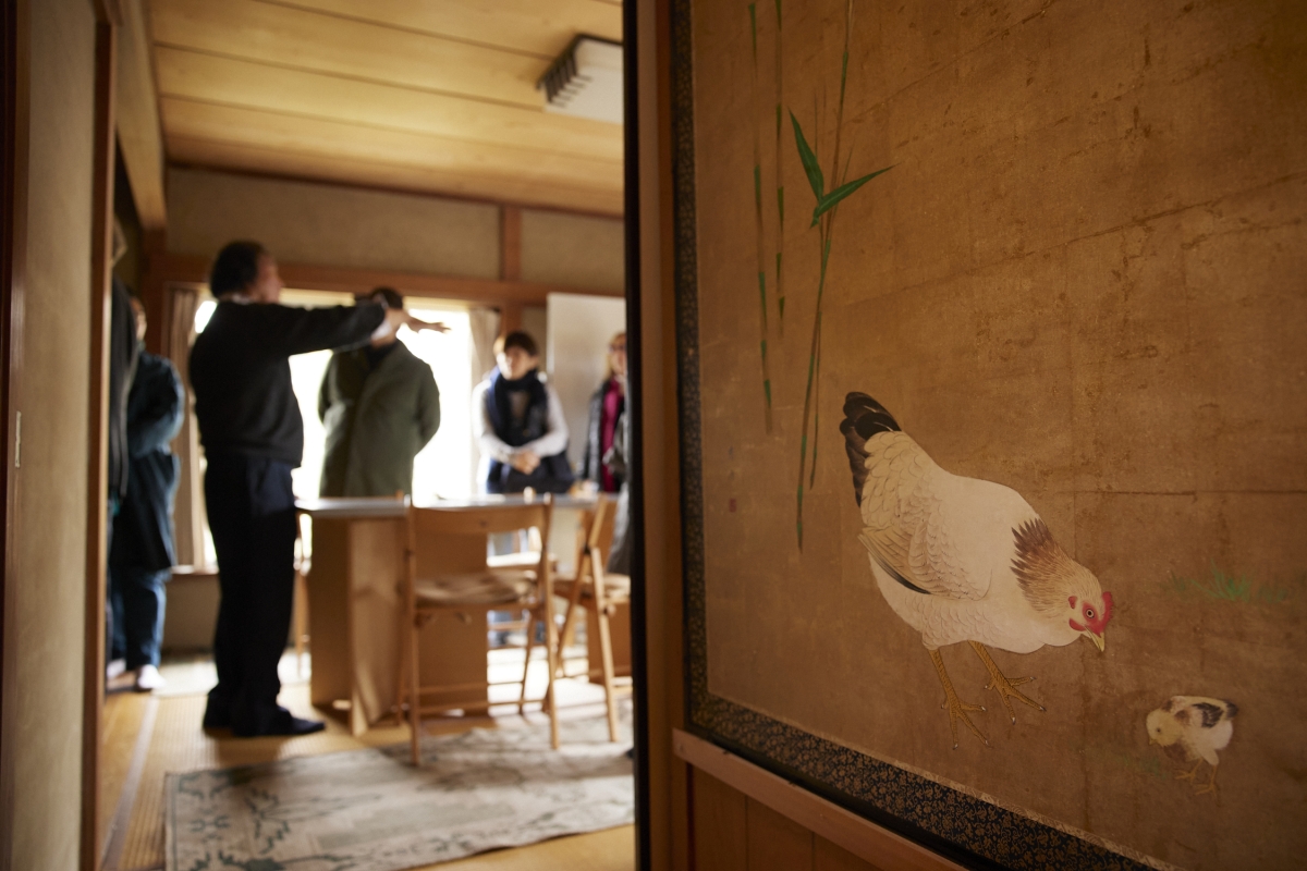 Japanese traditional painting of a chicken and a chic on the right wall overlooking Mr. Takishita with the visitor in the far room