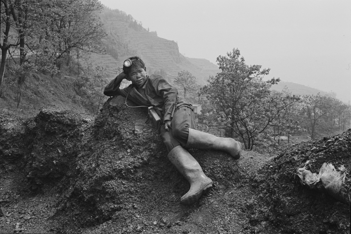 Black-and-white photograph of a boy approximately 11 years old lies on his side on a mount of dirt in a rocky landscape. He is wearing miner's clothes: coveralls, boots, and a headlalmp.  