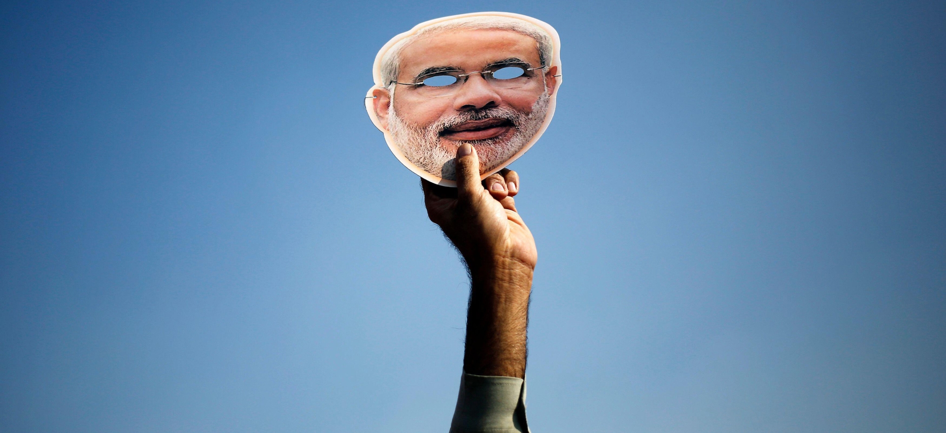 A hand holds up a Narendra Modi mask in front of a blue sky.