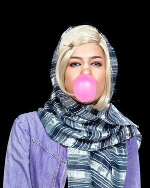 A woman wearing a purple denim jacket, striped headscarf and butterfly hair pin stares directly out at the viewer. She has blonde hair and blue eyes and a bandage on the bridge of her nose. She is blowing a bubble with bright pink bubble gum. 