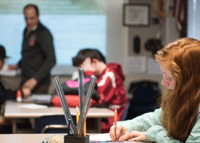 A student works with her class and teacher in the background.