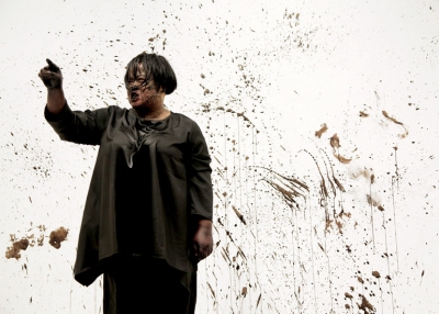 Performance artist Melati Suryodarmo stands covered in black ink with her right finger pointed upwards. 