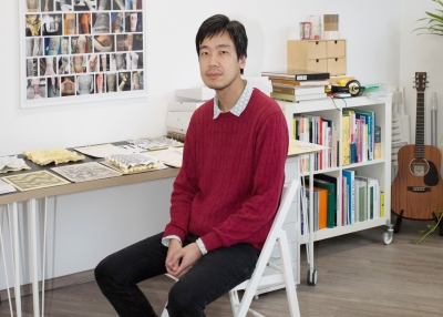 A man wearing a red knit sweater sits in front of a table displaying various prints and artwork.