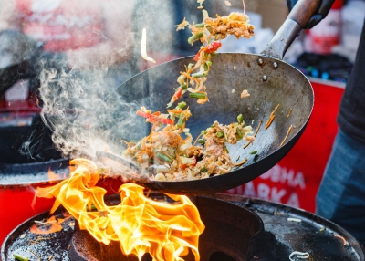 dynamic picture of a wok in motion, stir-frying colourful vegetables and meat. There's a bright flame contrasting the colour of the wok; thick smoke partially covering the wok and the dish, hinting at the rich smoked flavour the dish will have 