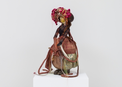 A doll-like figure is dressed in a brown and pink fitted jacket, full skirt of red, gold, and green and a large brown hat with pink flowers and white and orange ribbon and thread. The figure appears to be a woman and is holding what looks like an elephant tusk. She stands atop dark, brassy colored beast-like feet. 