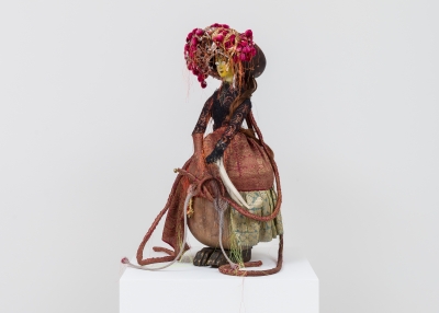 A doll-like figure is dressed in a brown and pink fitted jacket, full skirt of red, gold, and green and a large brown hat with pink flowers and white and orange ribbon and thread. The figure appears to be a woman and is holding what looks like an elephant tusk. She stands atop dark, brassy colored beast-like feet.