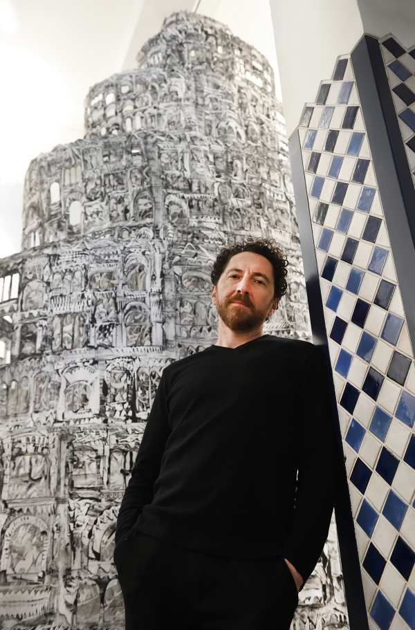 A man with a beard dressed in black stands in front of an elaborate drawing of a tower