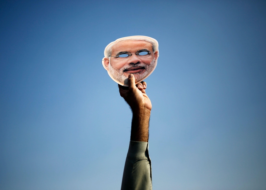 A hand holds up a Narendra Modi mask in front of a blue sky.