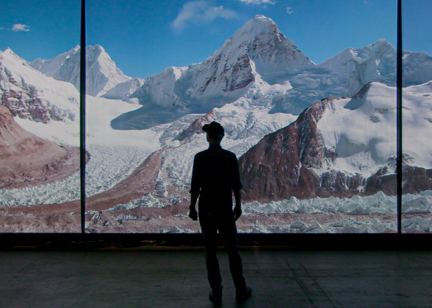 A person stands in front of a three-screen panoramic view of the Himalayas in full color.
