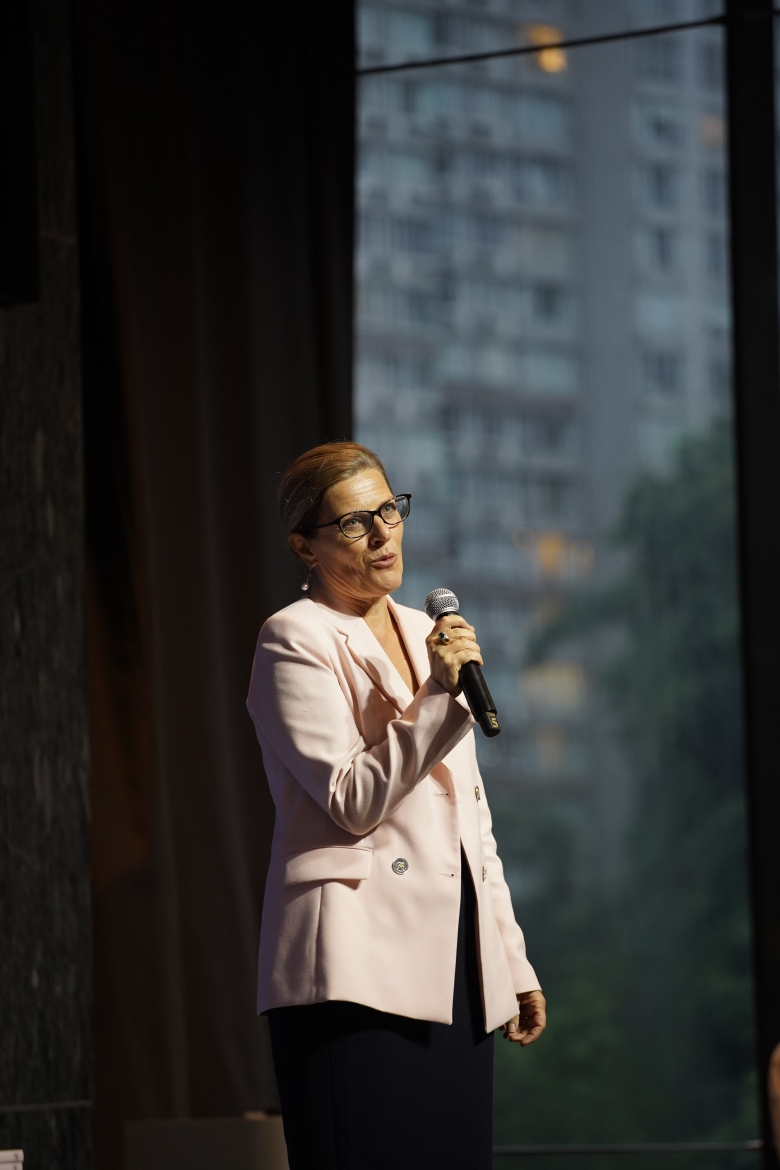A portrait image of one of the consul generals wearing a pink blazer with glasses holding a mic and making her speech.