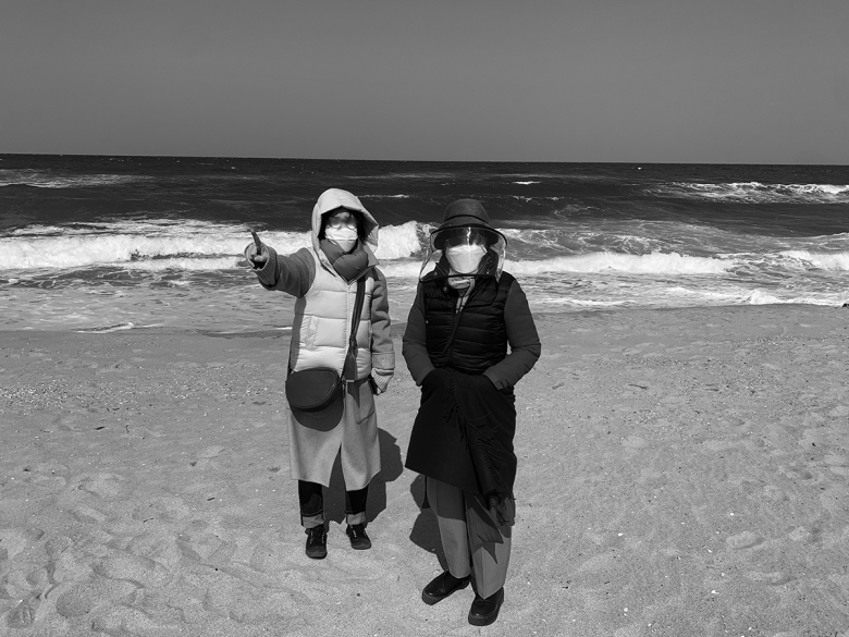 Two figures stand on the beach wearing facemasks and coats