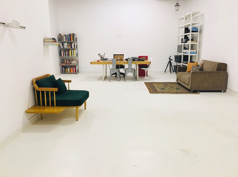 A room with white floor and walls. There are shelves of books, a green armchair, brown soda, and a wooden desk with a computer and office supplies. A variety of chairs are arranged around the table