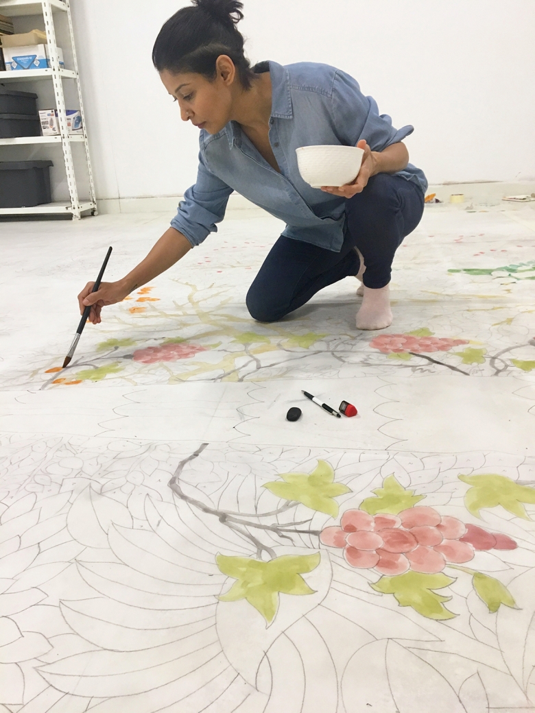 A person in a blue button-down top and dark pants crouches on top of a large drawing surface, holding a paintbrush and drawing patterns that look like grape vines