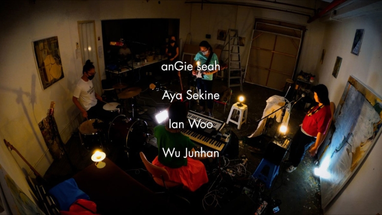 Five people are gathered in a music studio. Four are at instruments—drums, keyboard, guidar, and microphone—and a fifth stands watching. They wear masks and face each other in a circle formation.