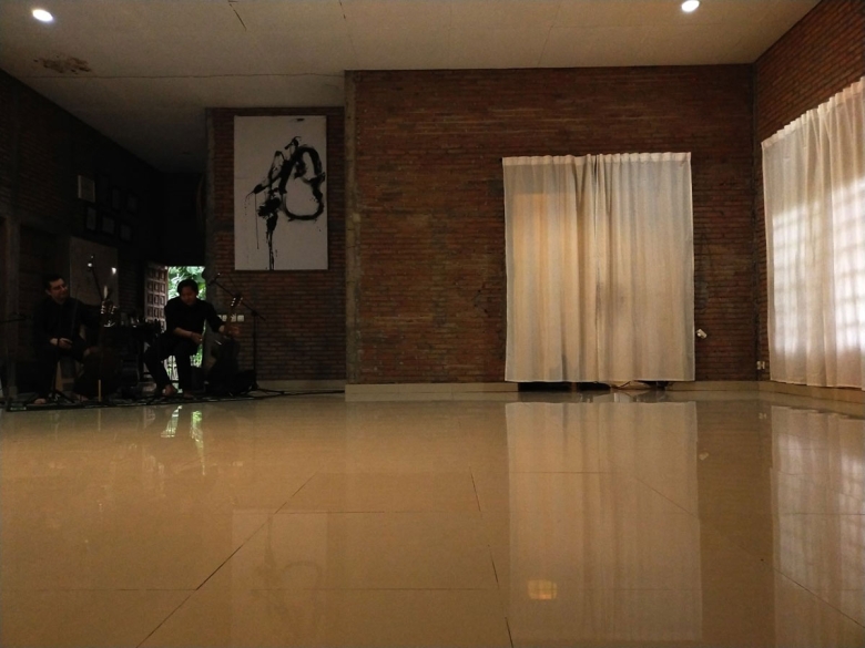 A studio interior with a linoleum, beige floor; brick walls; large, curtained windows, a rectangular white canvas with a black, expressive strock, and two people sitting in chairs near musical equipment.
