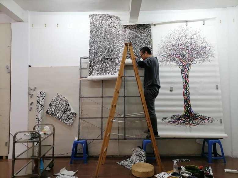 A photograph of the artist's studio. The artist appears on a ladder wearing a grey long-sleeved top and grey pants. A large, vertical sheet of paper hangs from scaffolding; it features an intricately drawn tree with hundreds of interlaced branches and roots drawn in black, red, blue, yellow, and green. A cart, stools, rolls of paper, paints, and bowls are visible in the room.